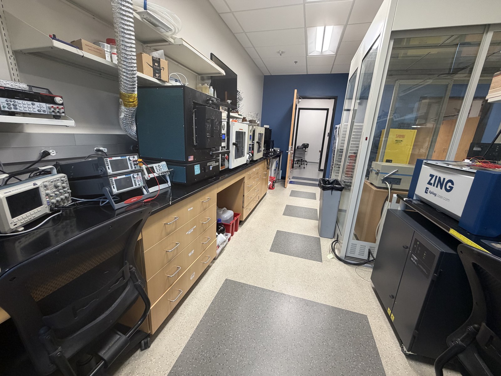 Lab bench lined with oscilloscopes, multimeters, an oven, and other electrical characterization equipment, with an Epilog Zing laser cutter and flammable storage cabinet down the hall.
