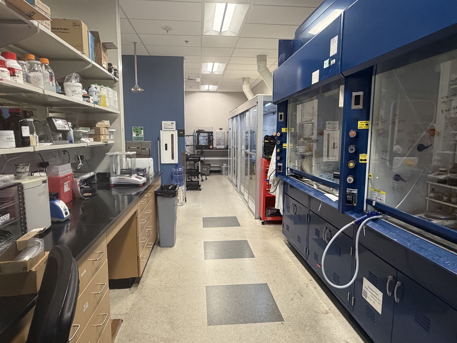 Shelving with chemical reagents next to an analytical balance, with a chemical fume hood and additional lab equipment down the hallway.