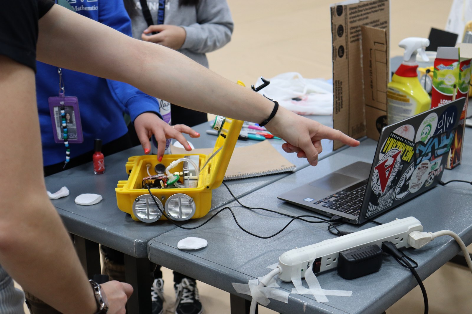 A student points to a laptop screen next to an open lab-built environmental sensor on a classroom table.